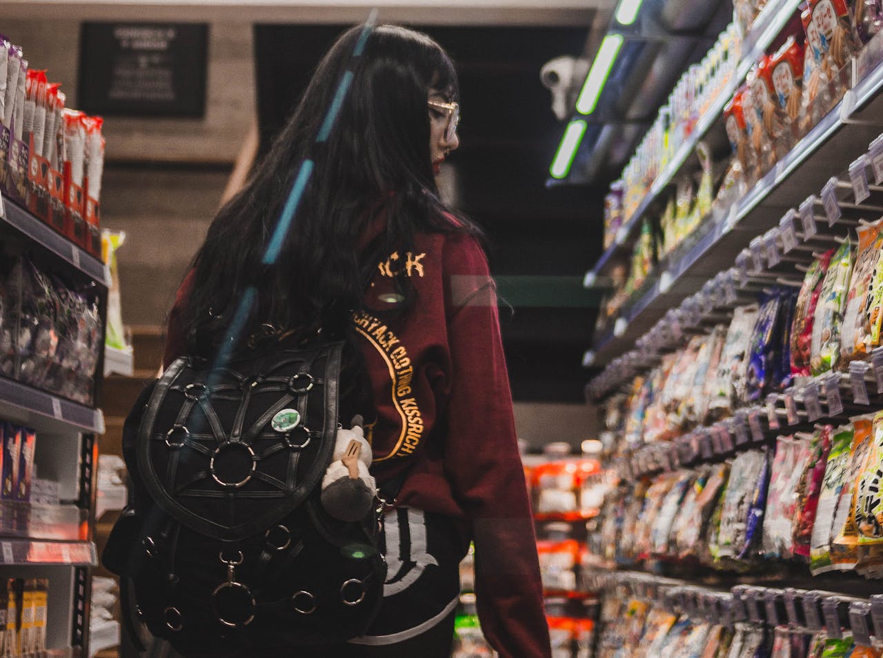 Woman seen from behind shopping in a snack aisle, wearing glasses and a fashionable backpack.