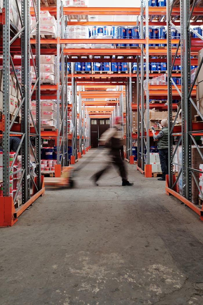 Blurred motion of a man walking in a spacious warehouse aisle between tall shelves.